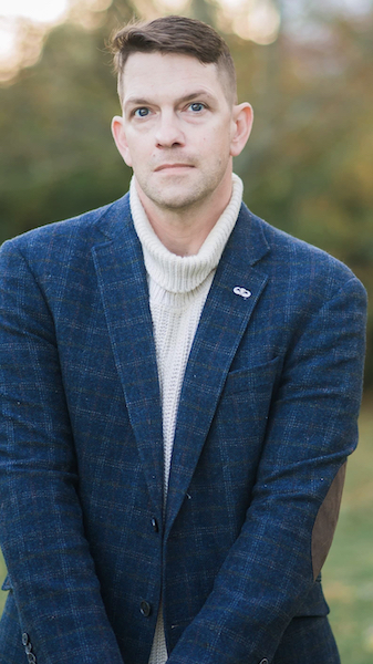 Anthony Roberts, tall Greek white man with brown hair, dark blue eyes, slight scruff, wearing a white turtle neck sweater and a blue tweed jacket standing infant of blurred out green trees and sunlight.